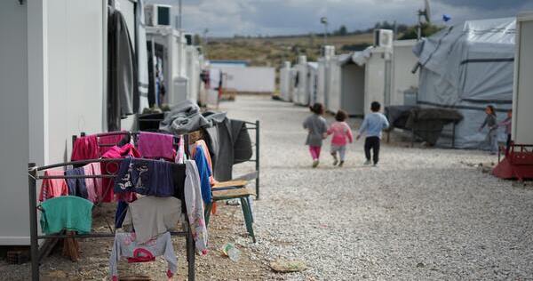 This Unsplash.com photo, by Julie Ricard (CC0 Licensing) depicts an everyday scene in a Syrian refugee camp in Greece, with three children in the background walking down a gravel road between refugee tent-housing, while in the foreground we see clothes hanging on makeshift lines outside one tent, to dry.