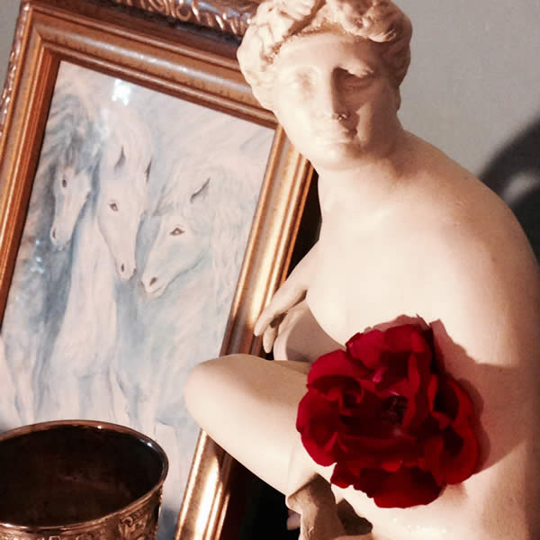 a statue of Venus holds a paper rose and sits before a painting of three, white horses