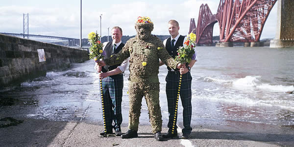 two men holding flowers stand beside a third dressed as the Burry Man