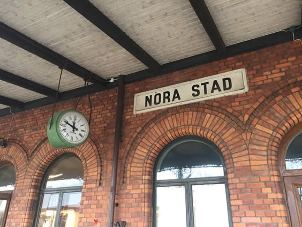 a brick building showing an exterior clock with the Swedish phrase "Nora Stad" (translation: Nora Station)