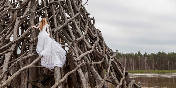a woman in a long white gown standing on a conical structure made of tree limbs in a tidal basin