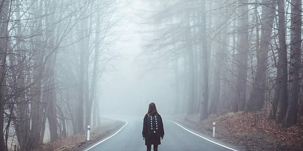 a woman standing in the middle of a road facing away from the camera looking into a fog bank