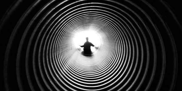 a person stands at the end of a long, ribbed pipe backlit by the sun