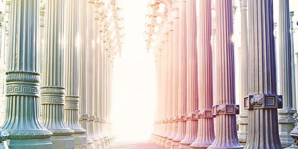 a photograph taken from within a building showing a series of columns as the sun shines into the hallway formed by them