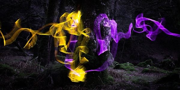 a digially manipulated photo of a black-and-white tree trunk with swirls of irridescent yellow and purple smoke surrounding and spreading away from it