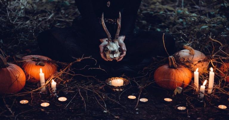 A person is holding an animal skull near the ground where pumpkins, tapers, and tealights lay.