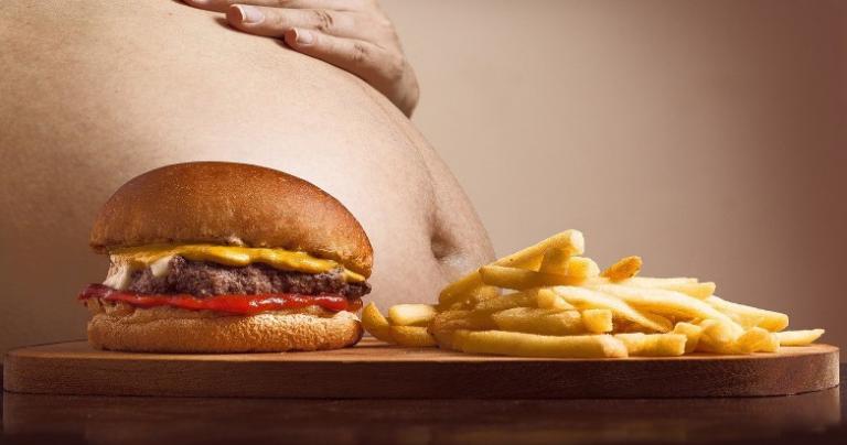 A person placing a hand on their bloated stomach with a hamburger and fries in the foreground.