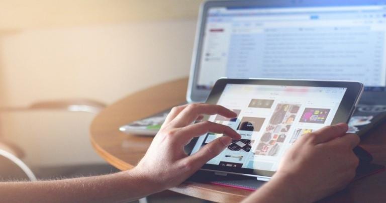 Hands holding a tablet with a computer in the background.