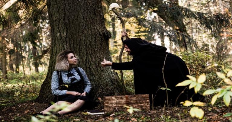 Woman sitting under a tree being visited by a stranger in a dark hooded robe and holding a staff.