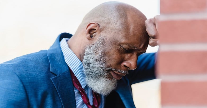 Discouraged bald black man in suit and loosened tie leaning against a wall