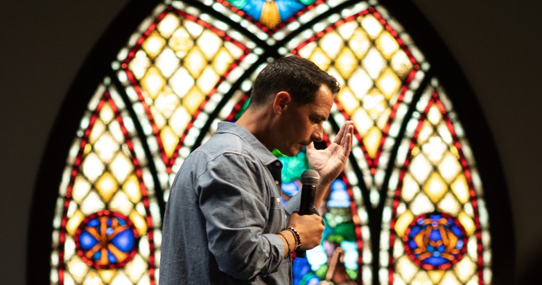 white male preacher in front of stained glass window