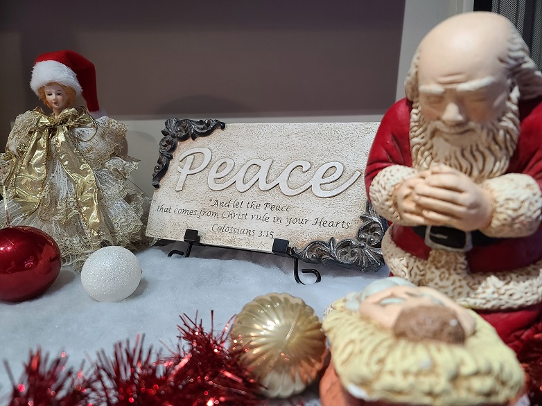 Santa kneeling before Jesus in manger while angel looks on, wearing Santa's hat