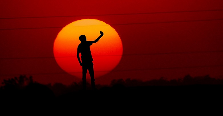 Man in profile, taking selfie, with huge orange sun behind. 