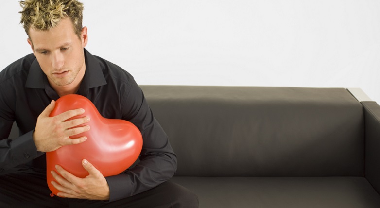 Man in black sitting on sofa, sadly holding red heart balloon