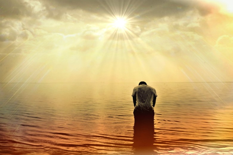 Find Peace Through One-Word Prayer. Man kneeling in water, facing away from viewer, praying towards sun in clouds
