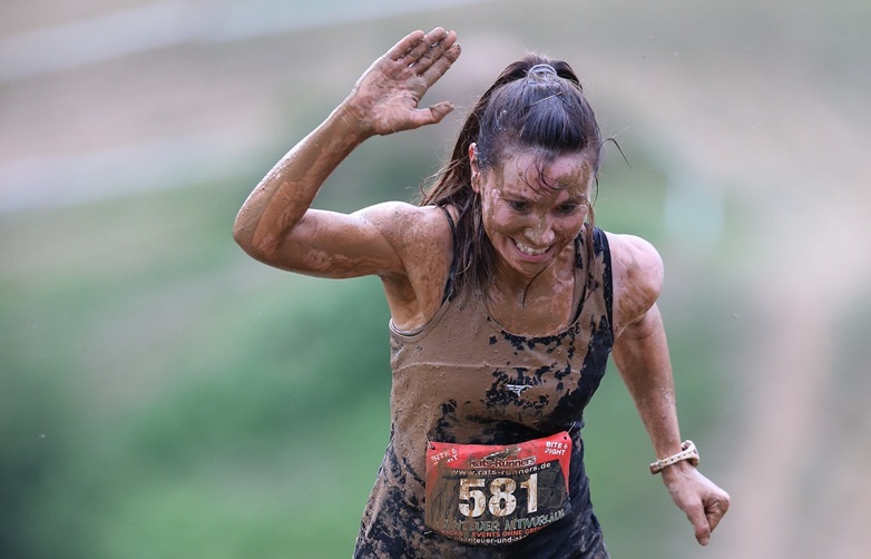 Muddy woman running in steeplechase race, raising one hand and smiling