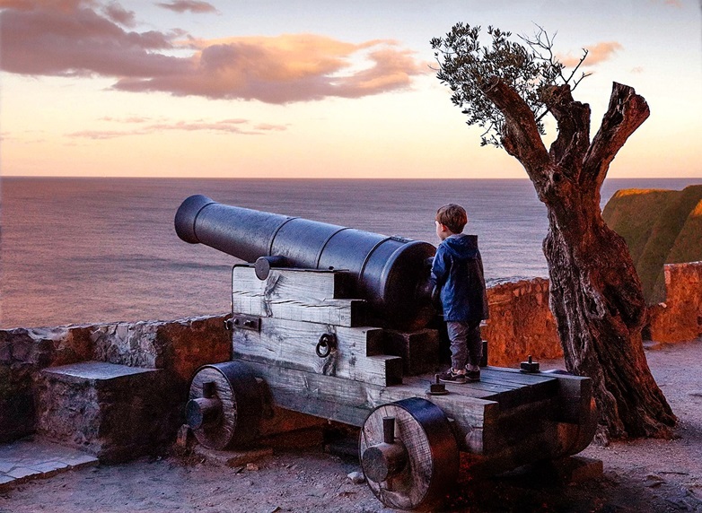 Young boy climbing on a cannon, overlooking the ocean.