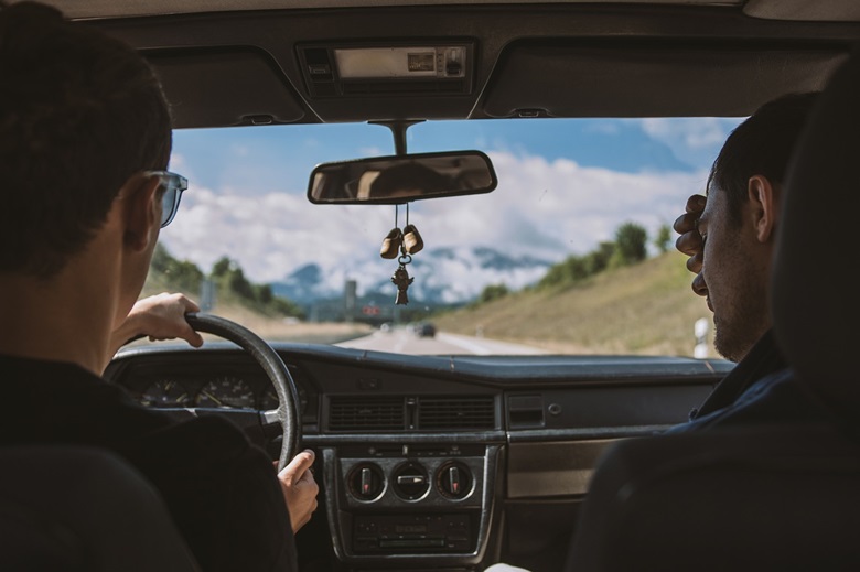 Two men in front seat of vehicle. Car is driving down highway with mountains in the distance, seen through windshield