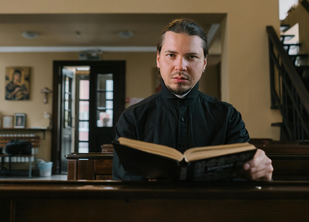 Pastor sitting in pew, holding a large Bible, looking into camera lens