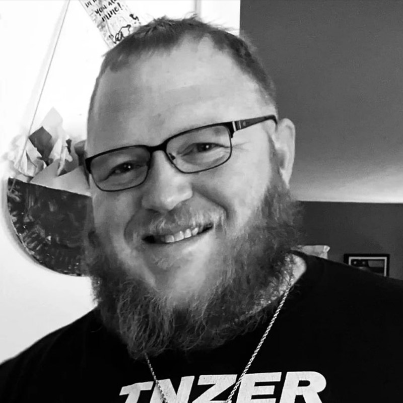 Black and white photo of a strikingly handsome and humble bearded man. He smiles while wearing an Inzer powerlifting shirt and a necklace with a miraculous medal, St. Michael medal and a cross made of nails.
