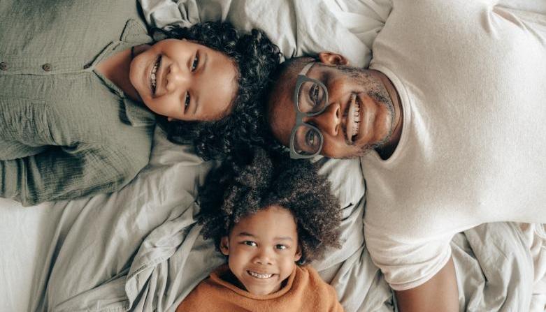 A father lays on his back with two of his children laying on their backs. Their heads meet in the middle, looking up at the camera. They all smile broadly.