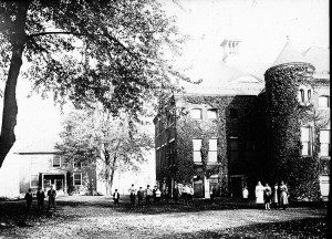 old black and white film photo of an old brick building to the right of the frame. small people in white stand in front of it. a large tree hangs over the top left half of the frame. 