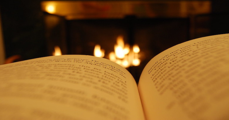 A book lies open in the darkness of a room with a fireplace in the background.