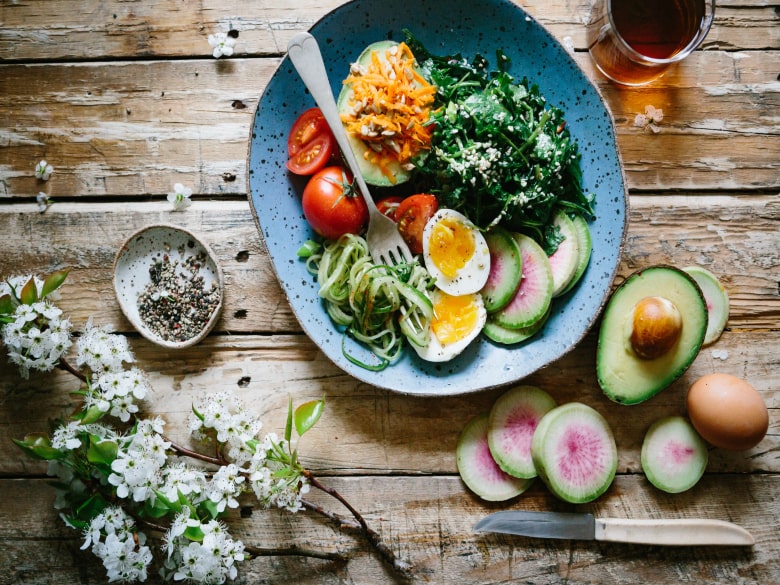 Fresh tomatoes, zucchini noodles, poached eggs, radishes, carrots, and kale in a bowl drizzled with sesame oil and coconut aminos