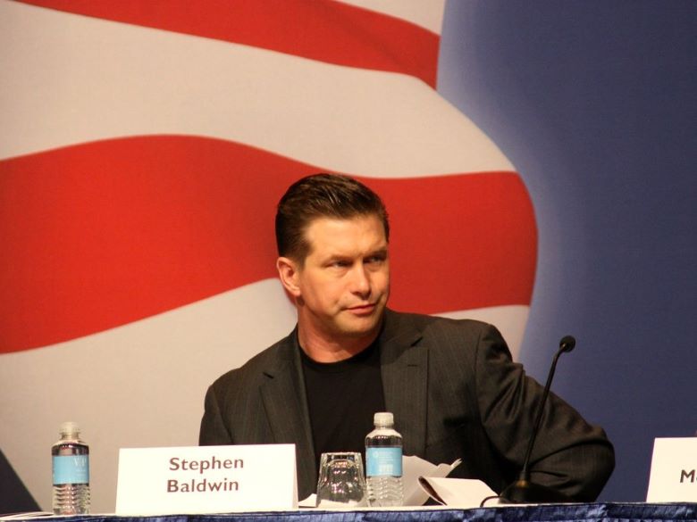 Stephen Baldwin seated on a panel with a nametag and water bottle in front of him and an American flag in the background