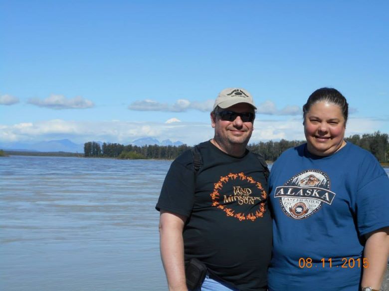 Husband and wife in t-shirts posing on the right with Talkeetnat River behind them and the peak of Denali visible in the distance behind a passing cloud.