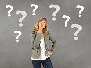 A woman stands in front of a grey background with her hand on her chin as if pondering. There are question marks all around her head.