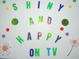 Poster with words "Shiny and Happy on TV"; smiley faces and daisies