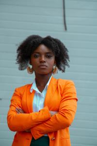 Black female wearing white shirt under bright orange suit with large earrings; very serious facial expression, arms crossed