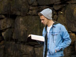 Young man in cap and jean jacket standing in front of rock wall, reading his Bible in peace