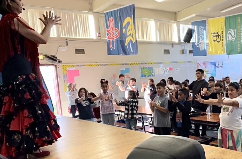 Woman in blue and red Flamenco dress teaching clapping to elementary students in school multipurpose room