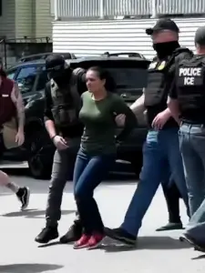 A distressed Brown-skinned woman is harshly led by two heavily-armed ICE police agents in body armor.