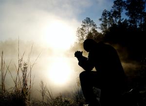 silhouette of man kneeling in prayer in front of a shadowy figure of light