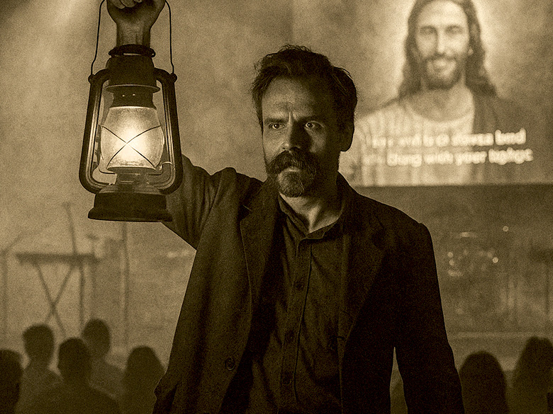 A sepia-toned image of a man holding a lit lantern aloft inside a fog-filled church. Behind him, a glowing image of Jesus is projected on a screen as the congregation sits in shadow.