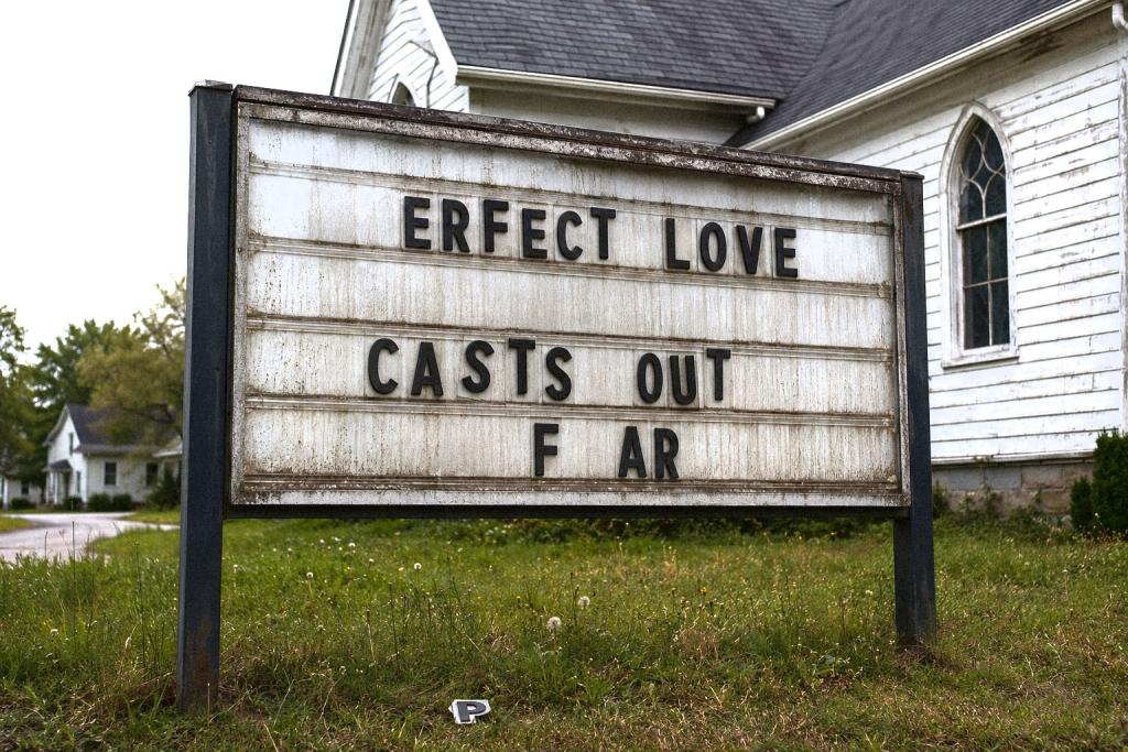 A weathered roadside church marquee with missing and crooked plastic letters partially reads “ERFECT LOVE CASTS OUT F AR” in front of an aging white clapboard church. Overgrown grass surrounds the base of the sign, and a fallen letter lies on the ground beneath it. The overcast sky and muted colors create a cold, uneasy atmosphere of suburban decay.