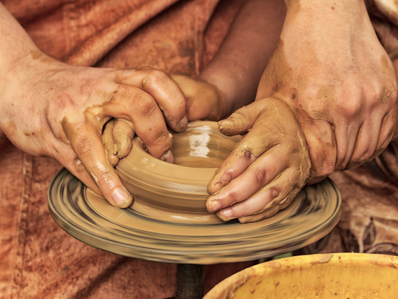 A clay bowl being made on a potters wheel with a set of older hands helping guide the hands of a child.