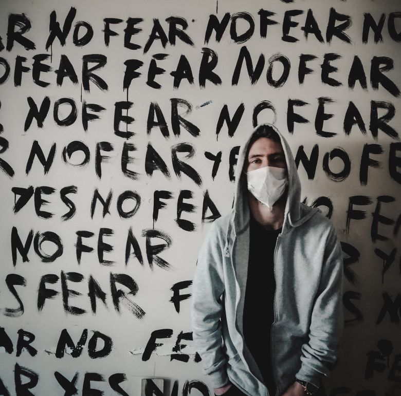 A man wears a hoodie and a face mask in front of a walk covered with "No Fear"