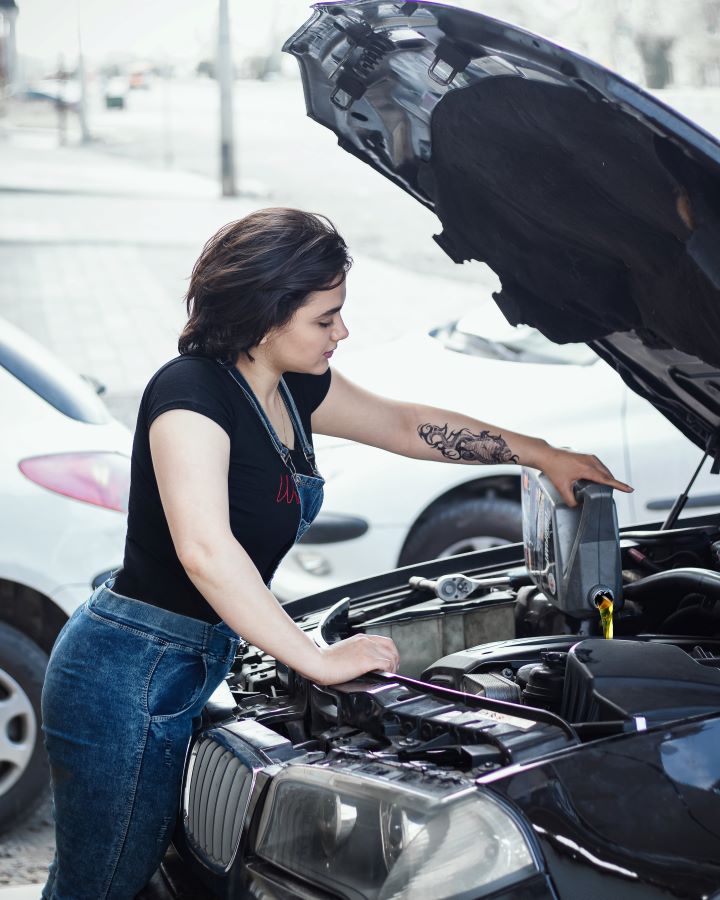 A woman with her car's hood up pours oil into it.