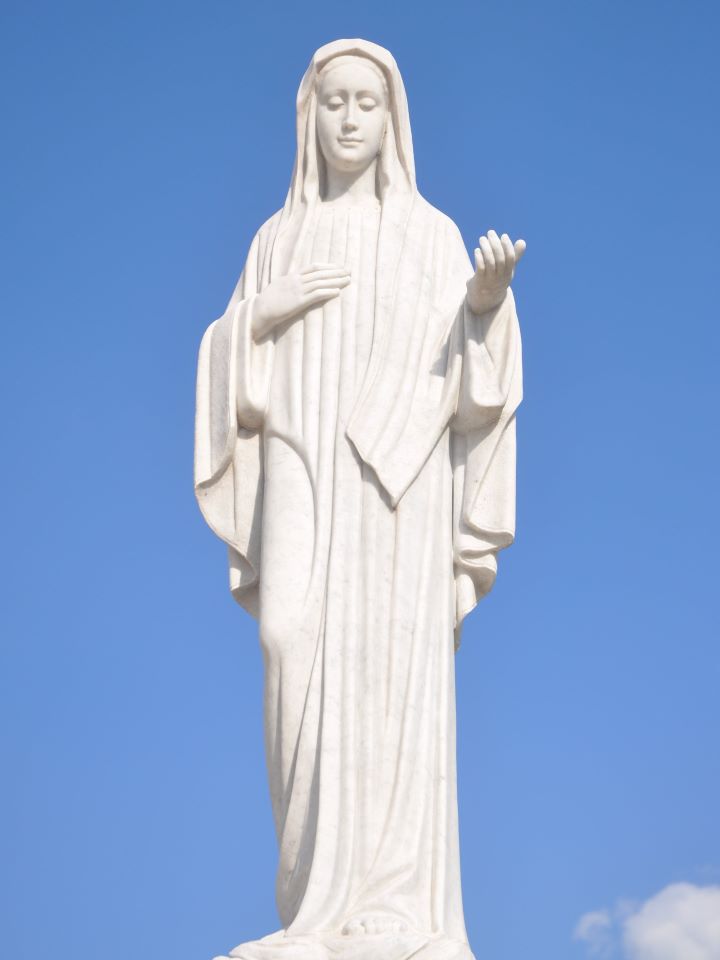 An all-white statue of the Virgin Mary with hand extended with blue sky behind her.