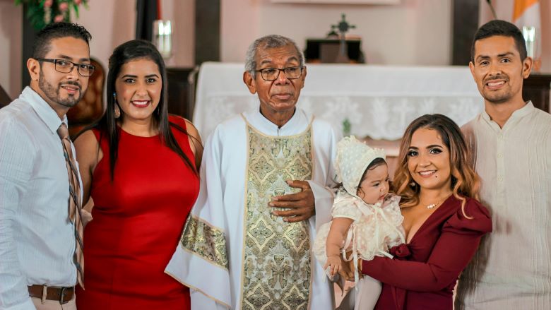 The photo depicts parents, godparents, a priest and a baby dressed in white.
