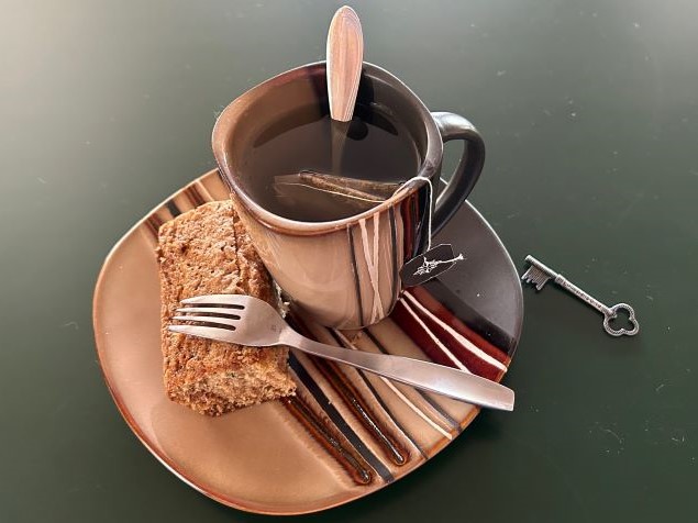 A cup of tea on a plate with a slice of banana bread and a fork. A key sitting next to the plate.