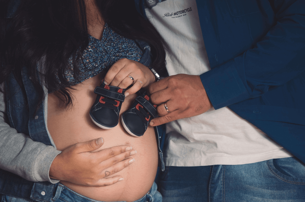 A pregnant couple taking a picture with baby shoes on an exposed expectant mother's belly.