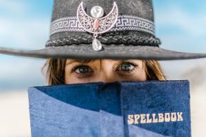 A blue spellbook held up to a white woman's face to just expose her blue eyes peering over it.