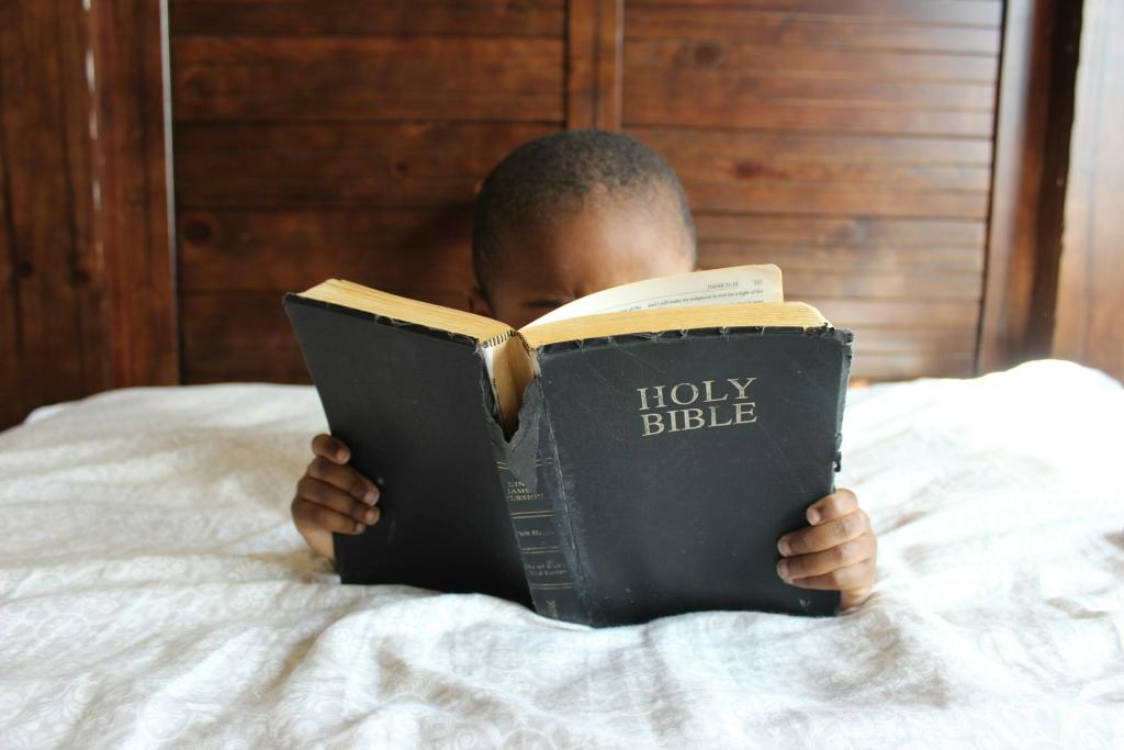 A young boy laying in bed reading a Bible, which is covering his face.