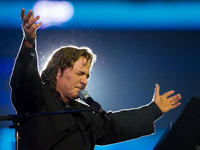 An image of a prophet named Kim Clement raising his hands to praise God in front of a blue background