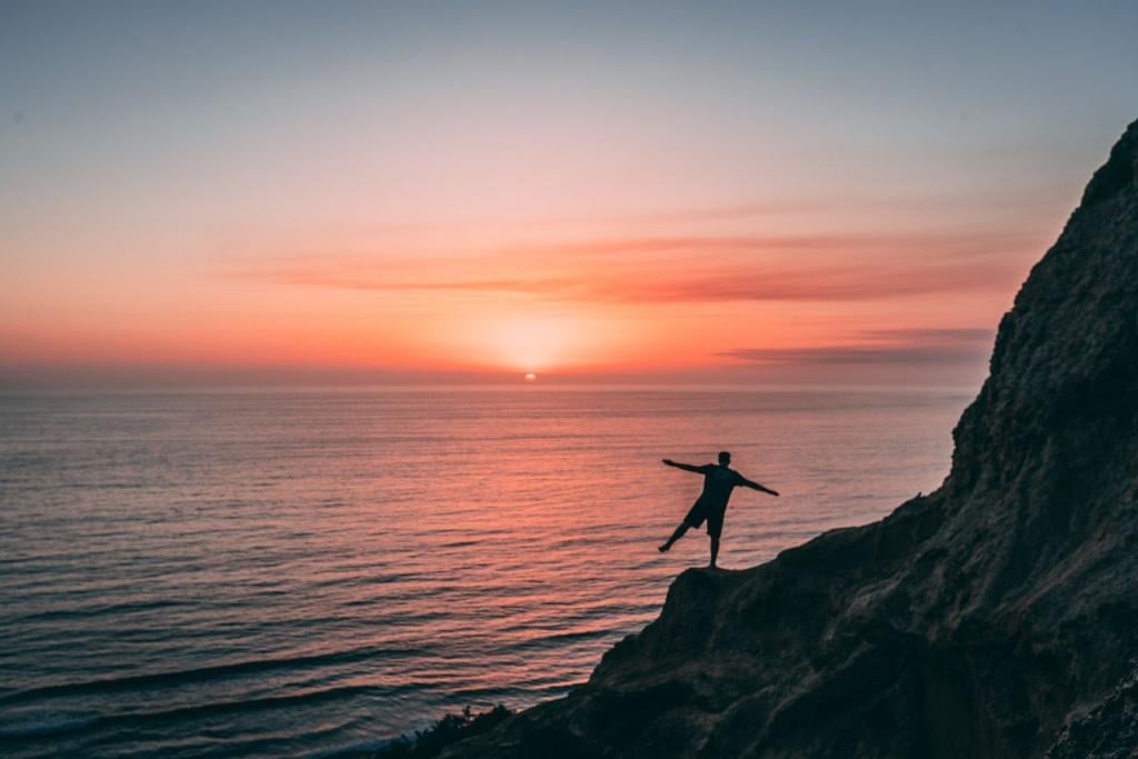 A man balances on a cliff staring in the sunset facing a horizon